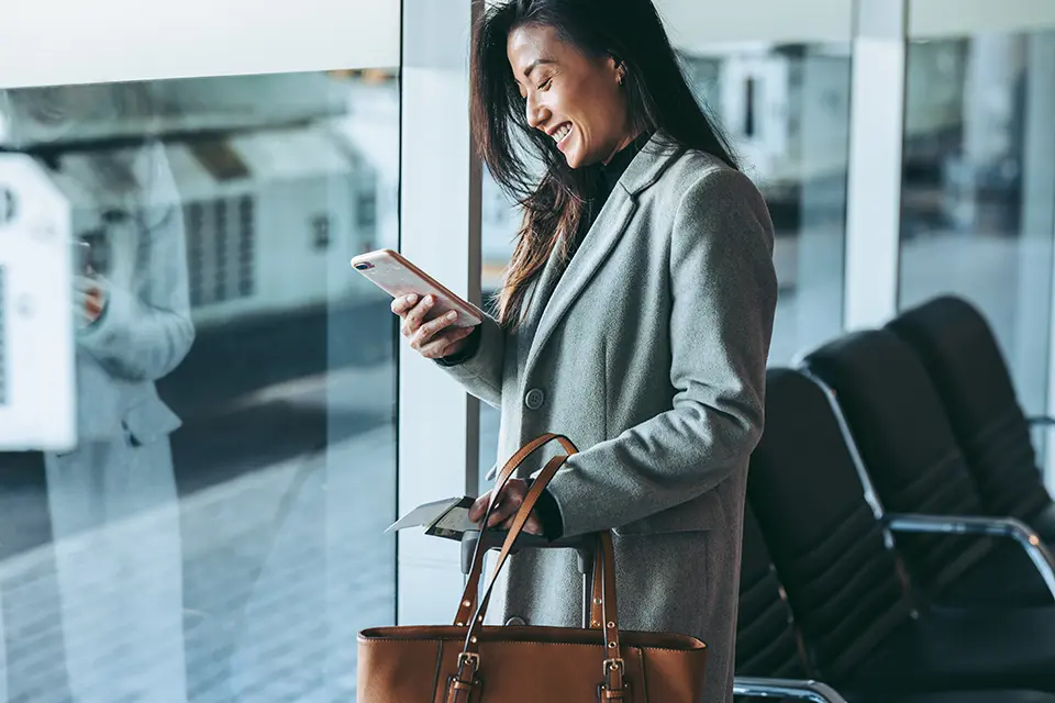 Woman holding a bag and texting on her mobile phone while waiting in an airport lounge, illustrating clients international travel.