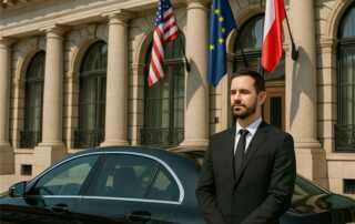 Chauffeur standing beside a luxury black sedan outside a government embassy building with flags.
